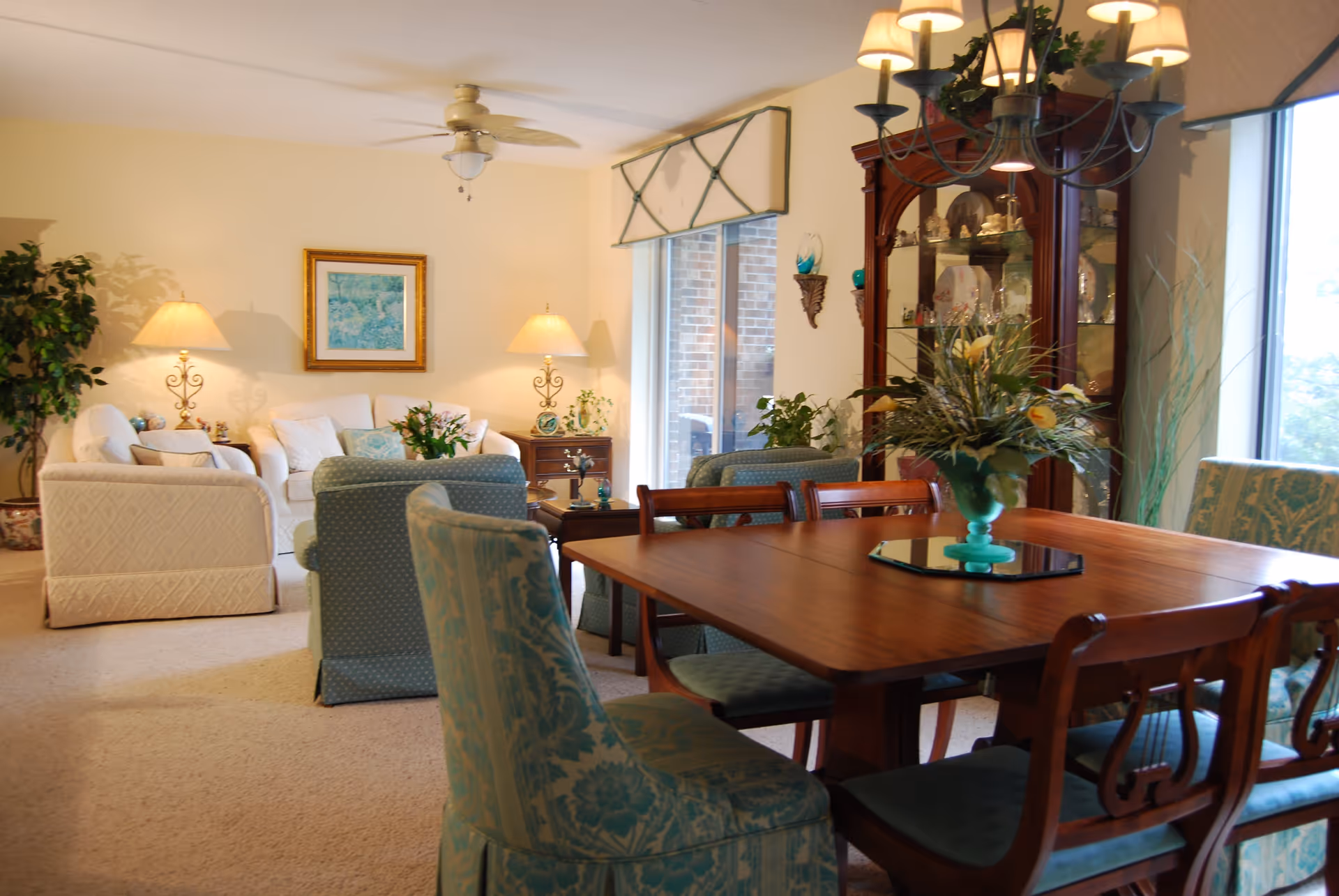 Bright combined dining and living area with a wooden dining table and upholstered chairs in the foreground and sofas, lamps, and a china cabinet in the background.