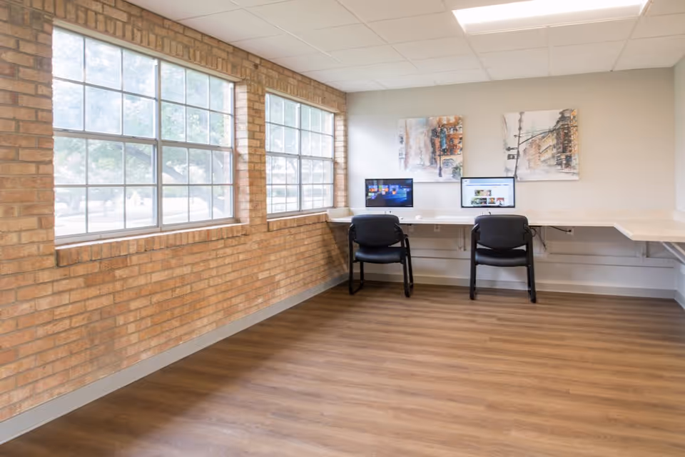 A bright room with large windows along one wall, featuring exposed brick and light wood flooring. Two black chairs are positioned at a long white desk with two computer monitors. Two abstract paintings hang on the wall above the desk.