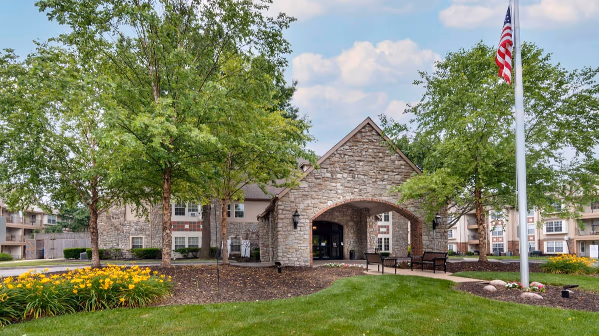 Stone-faced senior living building entrance with an arched porte-cochère, landscaped lawn, trees, benches, and an American flag.