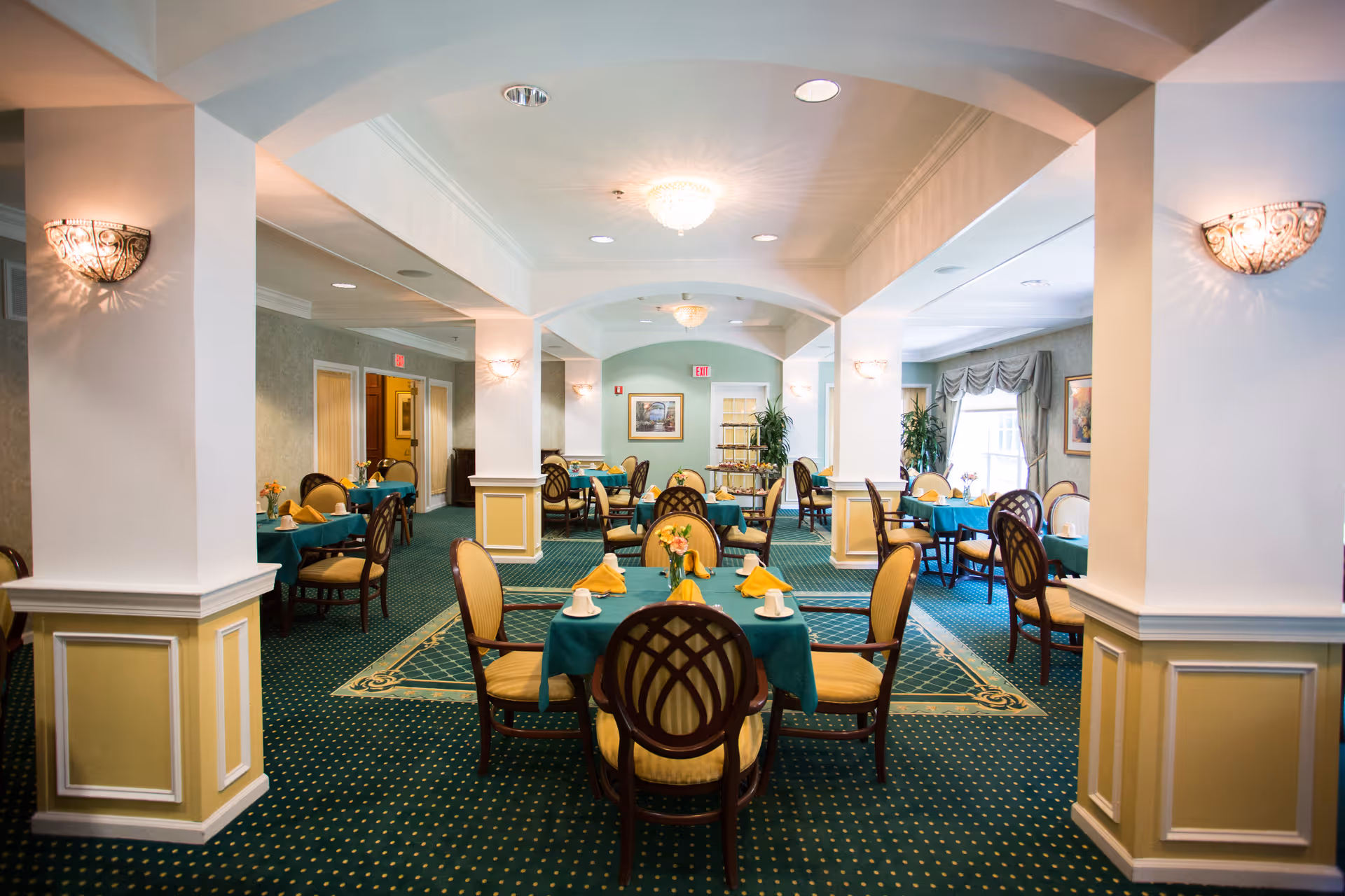 A well-lit dining room with multiple tables covered in teal tablecloths, each set with yellow napkins, cups, and small flower arrangements. The room features green carpet with a patterned border, cream-colored walls with wainscoting, decorative wall sconces, and large windows with drapes allowing natural light to enter.
