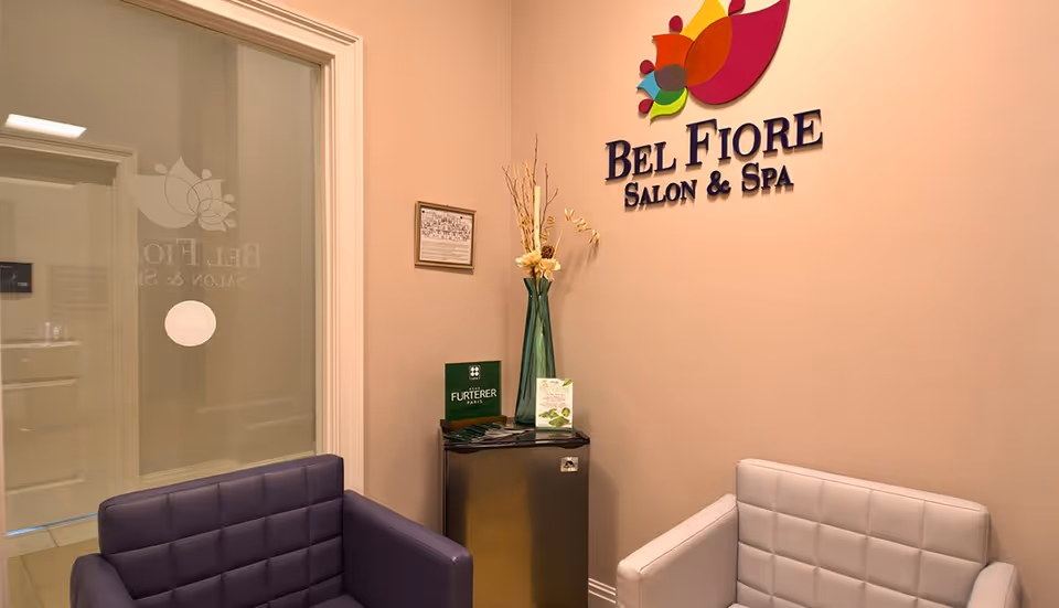 Interior view of a salon and spa waiting area with two cushioned chairs, one purple and one white, a small table with brochures and a tall vase with decorative dried flowers, and a wall sign that reads 'Bel Fiore Salon & Spa'.
