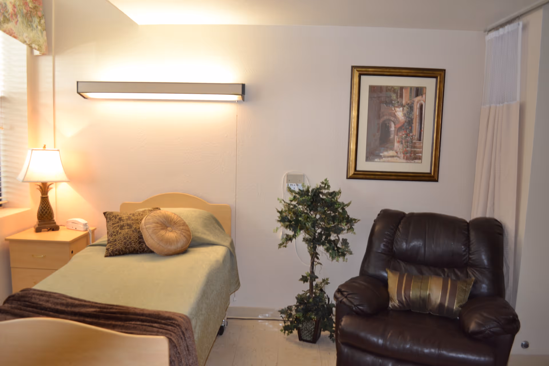 A small nursing facility bedroom with a single bed, bedside table and lamp, a potted plant, and a leather recliner.