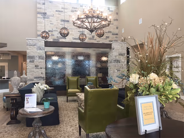 Lobby seating area with green armchairs, sofas, a stone water wall feature, chandelier, and a large floral arrangement.