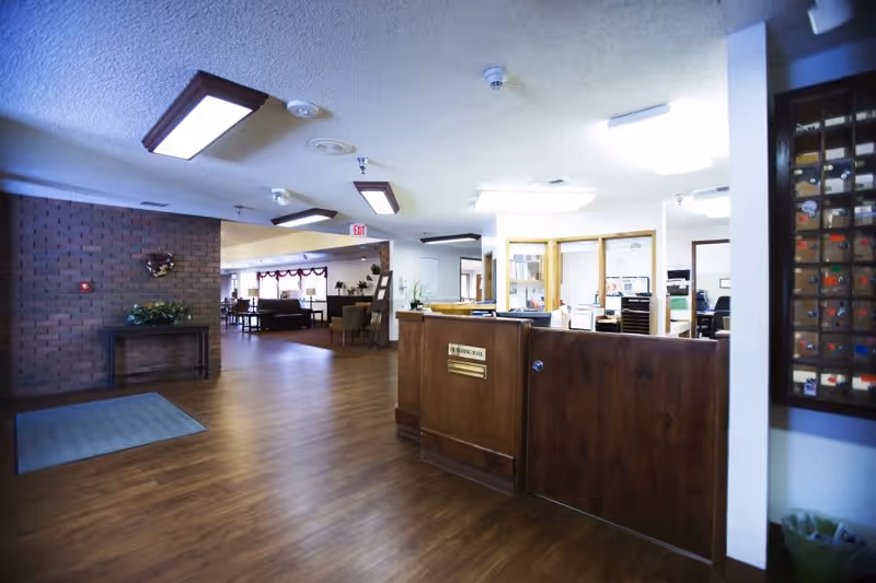 Interior view of a senior living facility lobby or reception area with a wooden reception desk, wood flooring, brick accent wall, and seating area with chairs and tables in the background.