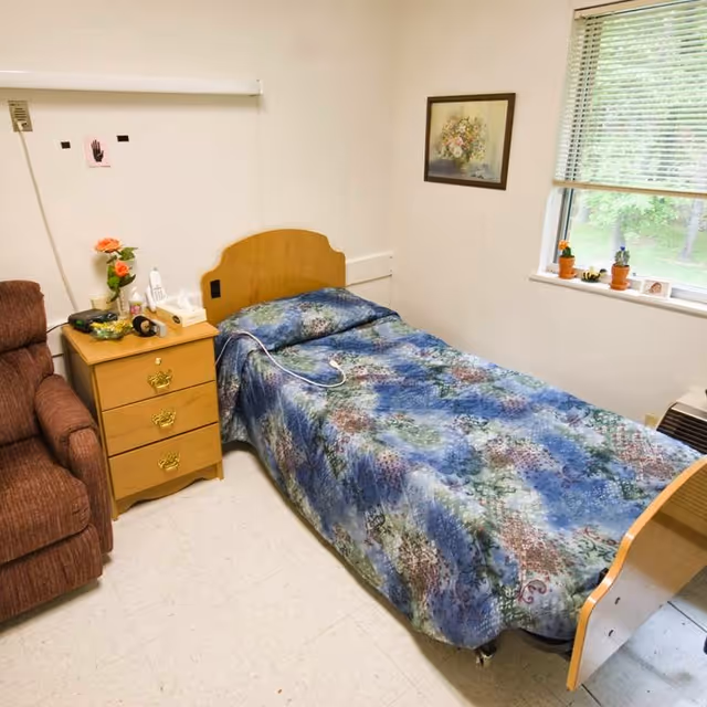 A small bedroom in a senior living facility with a single bed covered in a blue patterned bedspread. Next to the bed is a wooden nightstand with three drawers, holding a telephone, a box of tissues, a small vase with orange flowers, and some other small items. To the left of the nightstand is a brown upholstered armchair. On the wall above the bed is a framed floral painting. A window with blinds partially open shows greenery outside. The floor is tiled in a light color.