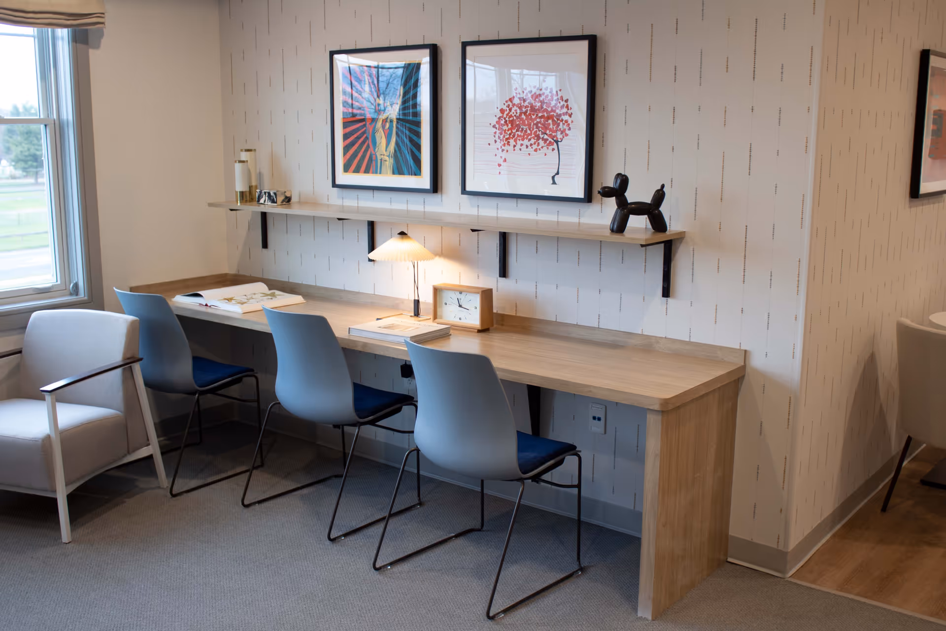 A modern workspace area with a long wooden desk against a patterned wall. Three blue chairs with black metal legs are placed at the desk. On the desk are an open book, a small table lamp, a clock, and some stacked books. Above the desk, a wooden shelf holds two framed artworks and a black balloon dog sculpture. To the left, there is a window with a beige Roman shade and a light-colored armchair with wooden armrests.