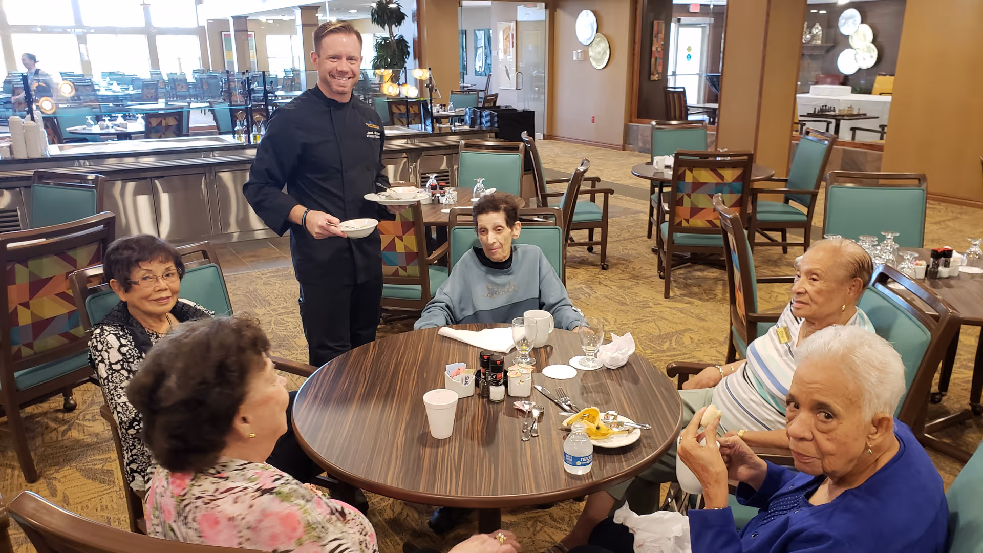 A group of older adults seated around a dining table with a server standing nearby in a retirement community dining room.