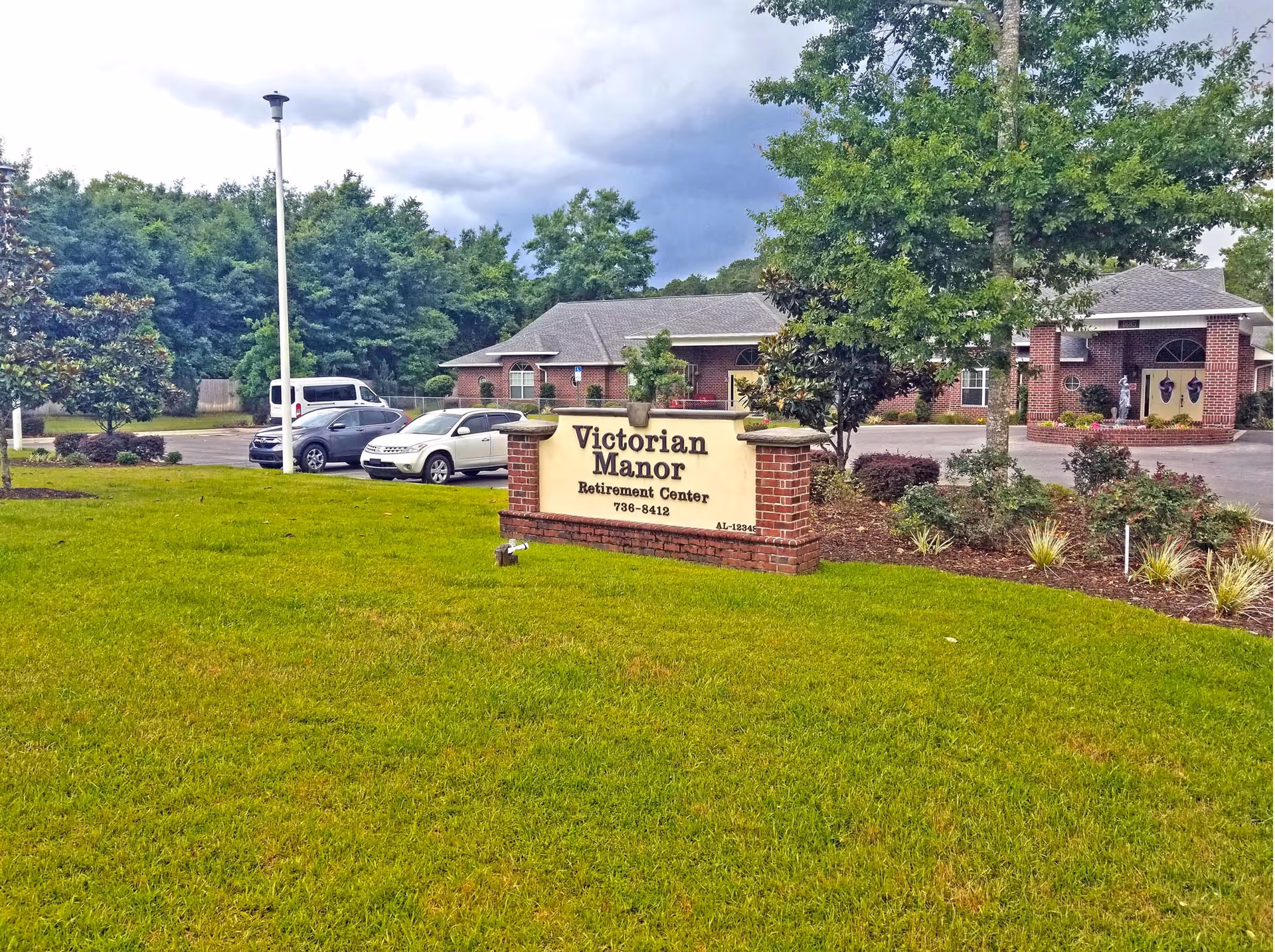 Entrance sign and front lawn of Victorian Manor retirement center with the brick main building and parked cars behind it.