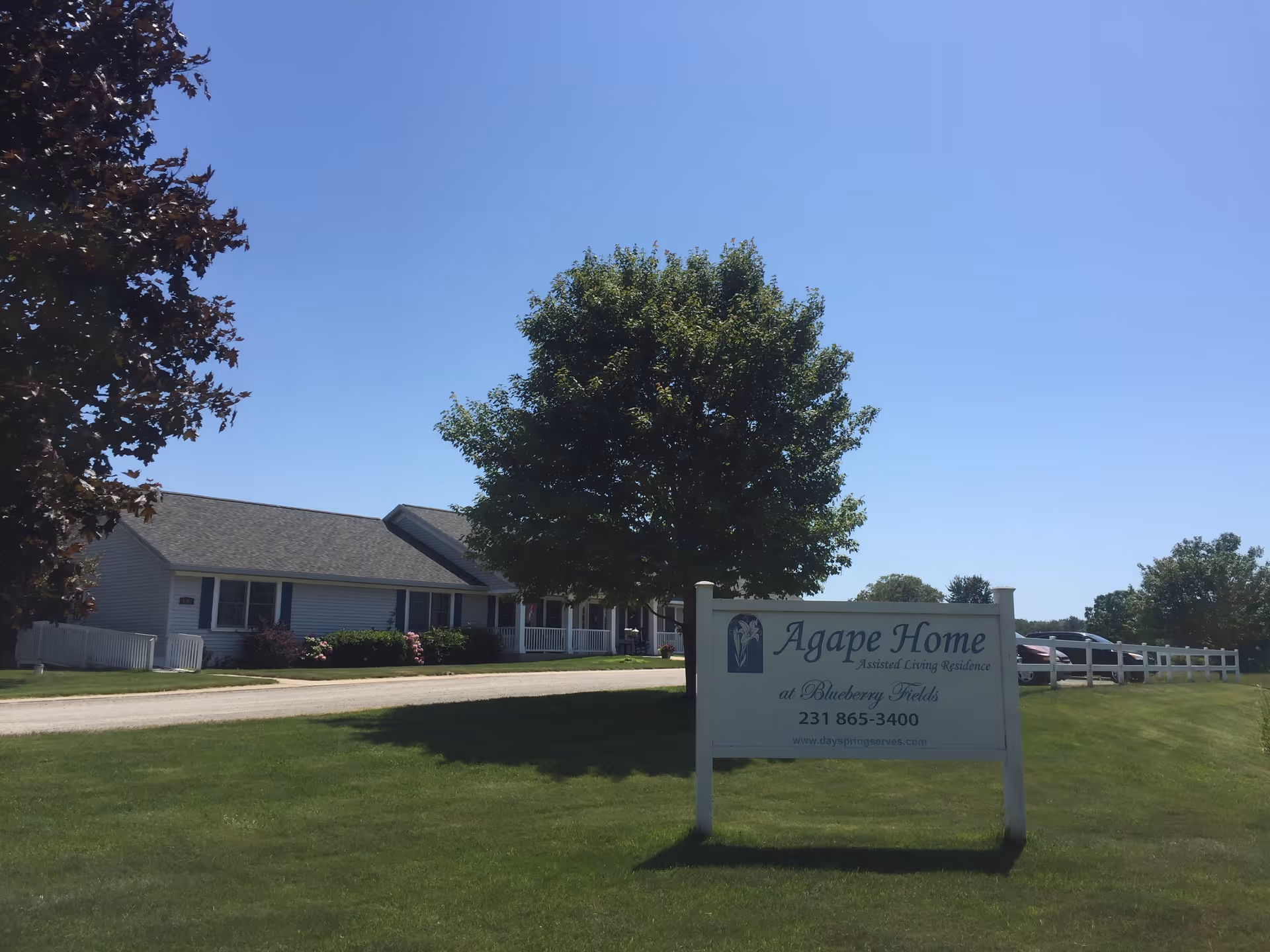Exterior view of Agape Home at Blueberry Fields, an assisted living residence, showing a single-story building with a pitched roof, surrounded by green grass and trees under a clear blue sky. A white sign with the facility's name, phone number, and website is prominently displayed in the foreground.