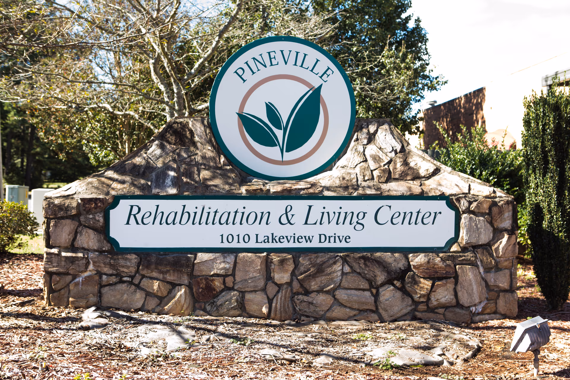 Stone sign for Pineville Rehabilitation & Living Center with the address 1010 Lakeview Drive, surrounded by trees and bushes under a clear sky.