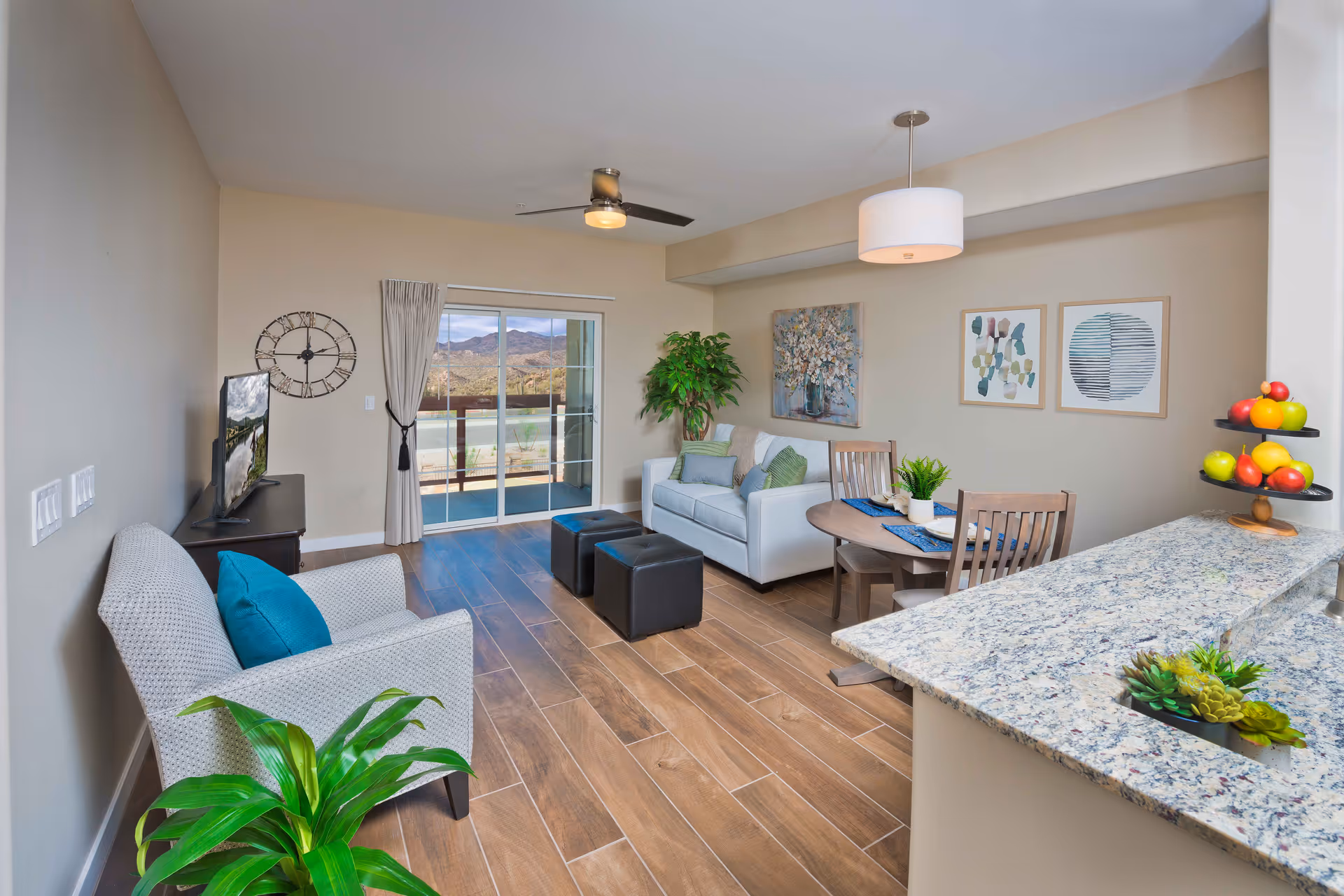 A bright and modern living room with wood-look tile flooring, a light gray sofa with green and blue cushions, a matching armchair with a blue pillow, two black ottomans, a round wooden dining table with four chairs, and a granite countertop with a tiered fruit stand. The room features a ceiling fan, three framed artworks on the wall, a large clock, a potted plant, and sliding glass doors leading to an outdoor patio with a view of mountains.