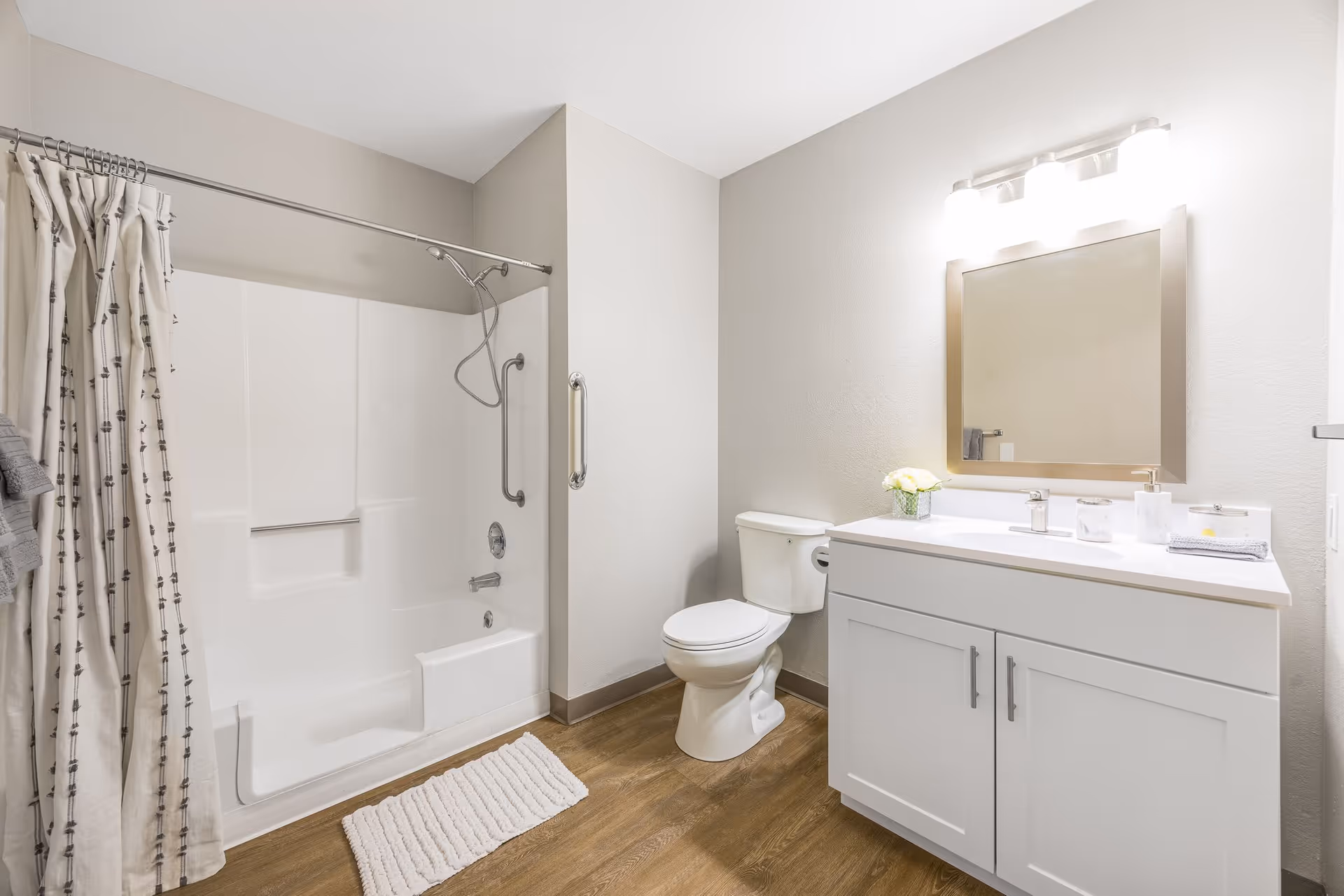 A clean and modern bathroom with a white bathtub and shower combination featuring a grab bar and a shower curtain with a subtle pattern. There is a white toilet next to a white vanity with a sink, a large mirror above it, and a light fixture with three bulbs. The floor has wood-like flooring and a white bath mat is placed in front of the tub.