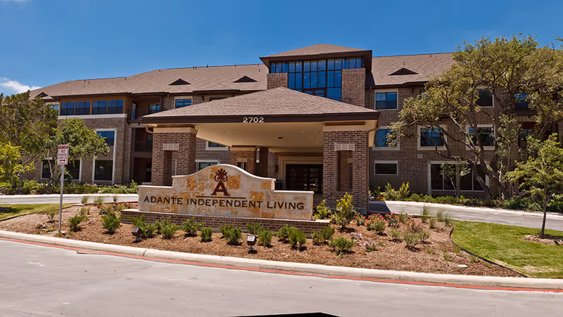Front exterior view of a multi-story brick building with a covered entrance and a large sign in front that reads 'Adante Independent Living'. The building is surrounded by landscaped greenery and trees under a clear blue sky.