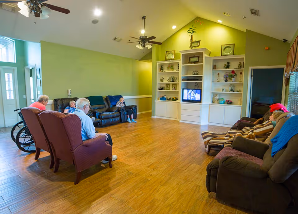 Several seniors seated in a bright communal living room with sofas, recliners, an entertainment center, and wood floors.