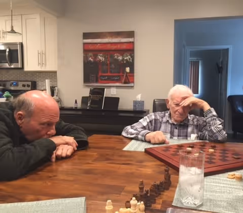 Two elderly men sitting at a wooden table in a kitchen or dining area, engaged in a game with chess and checkers pieces. One man is leaning forward resting his head on his arms, while the other man is looking thoughtfully at the board with his hand on his forehead. A glass of ice water is on the table, and kitchen cabinets and a painting are visible in the background.