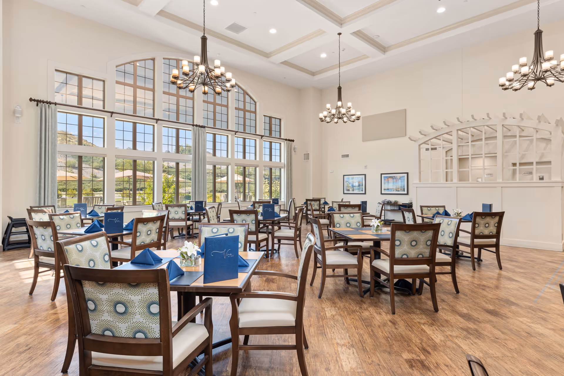 A spacious dining room with multiple wooden tables and chairs arranged neatly. Each table has blue napkins and menus, with small floral centerpieces. Large windows with curtains allow natural light to fill the room, and two chandeliers hang from the coffered ceiling. The room has light-colored walls and wood flooring, with framed artwork on one wall and an arched partition.
