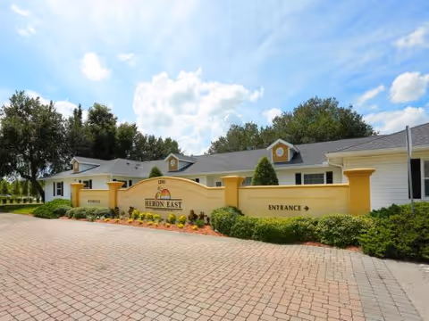 Exterior view of Heron East senior living facility showing a single-story building with white walls and yellow accents. There is a paved driveway in front, landscaped bushes, and a sign that reads 'Heron East' with an arrow indicating the entrance. The sky is partly cloudy with blue patches.