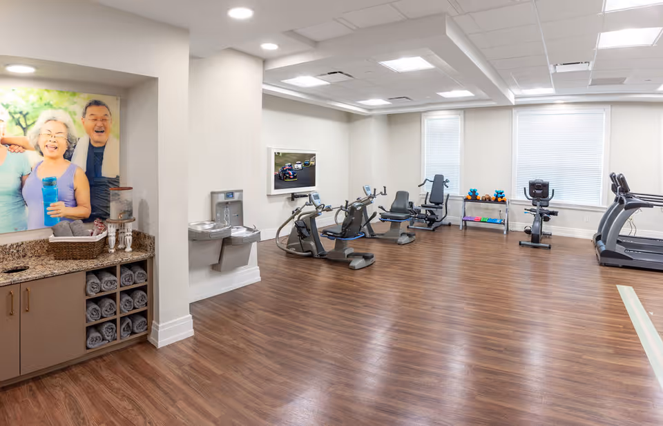 A fitness room with exercise equipment including stationary bikes, a treadmill, and weight racks. The room has wood flooring, white walls, and two large windows with blinds. On the left side, there is a counter with neatly rolled towels and a water dispenser, along with a large photo of smiling elderly people on the wall.