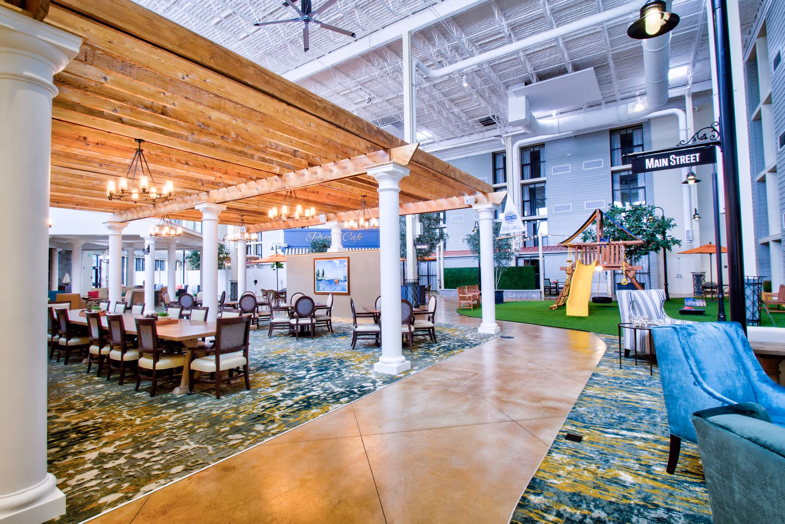 Indoor communal area with wooden pergola ceiling, chandeliers, multiple dining tables and chairs, a play area with a yellow slide, and seating with blue armchairs. The space has a high ceiling with exposed ducts and a sign reading 'Main Street'.