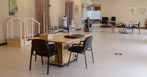 A spacious room with tables and chairs arranged for activities or therapy, including a wooden table with chairs, a set of stairs with handrails, exercise equipment, and various items on tables. The room has light-colored walls and floor, with artwork on the walls and curtains in the background.