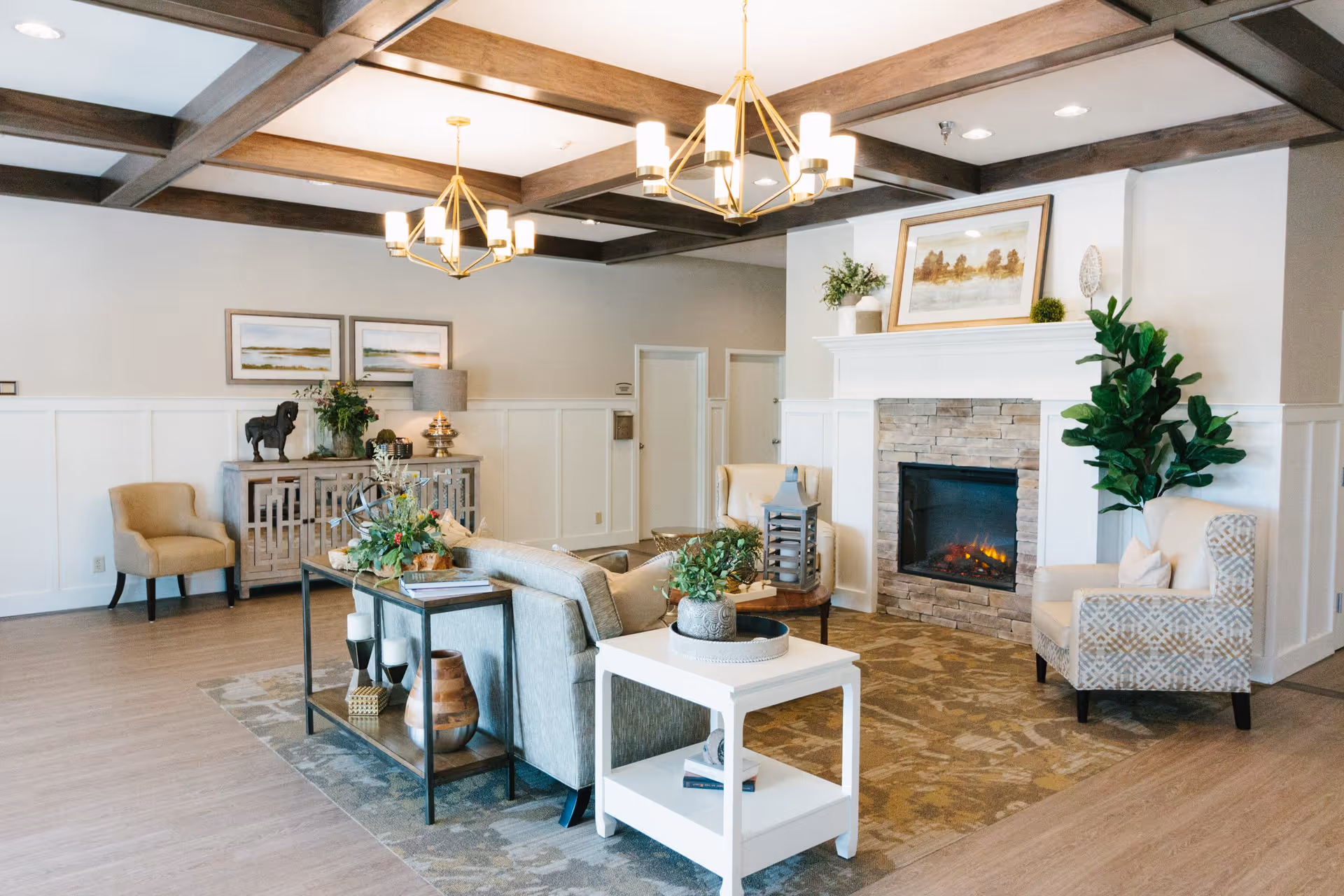 A cozy living room area with a stone fireplace, two armchairs, a sofa, and a coffee table. The room features wooden ceiling beams, two modern chandeliers, a large plant, framed artwork above the fireplace, and decorative items on side tables and a cabinet against the wall.
