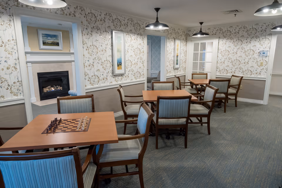 Dining-style common room with several wooden tables and upholstered chairs, a chessboard on a table, patterned wallpaper, pendant lights, and a fireplace visible through a doorway.