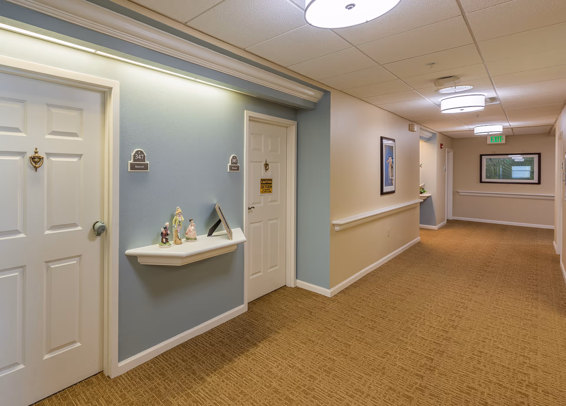 A well-lit hallway in a senior living facility with beige carpet and walls painted in light blue and cream. There are two white doors on the left side, one labeled 347 Reserved and the other 345 Home with a caution oxygen sign. A small white shelf between the doors holds decorative figurines. The hallway extends with framed artwork on the walls and ceiling lights illuminating the space.