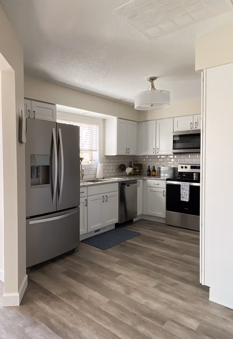 Modern kitchen with stainless steel refrigerator, dishwasher, oven, and microwave. White cabinets and subway tile backsplash with a window above the sink. Wood-look flooring and a ceiling light fixture.
