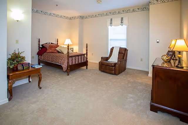 A spacious bedroom with beige carpet and cream-colored walls. The room features a wooden bed with a patterned bedspread and multiple pillows, a wooden side table with a lamp and books, a brown recliner chair with a white throw draped over it, and a wooden dresser with a lamp and decorative items. A window with blinds is visible behind the recliner chair.