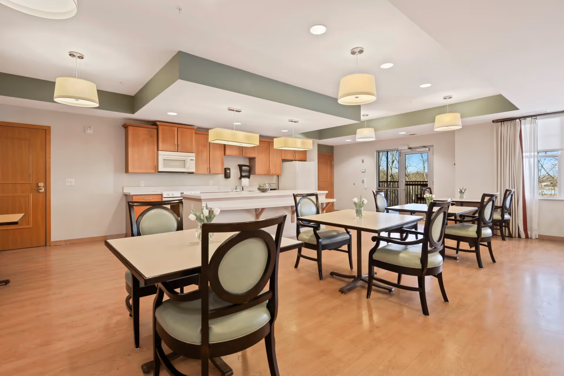 A bright dining area in a senior living facility featuring several square tables with green cushioned chairs. The room has light wood flooring, a kitchen area with wooden cabinets, a white refrigerator, microwave, and stove. Pendant lights hang from the ceiling, and large windows and a glass door allow natural light to enter the space.