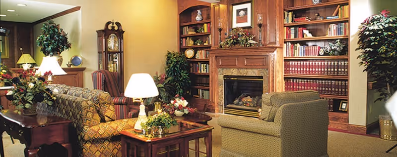 A cozy living room area in a senior living facility featuring a fireplace surrounded by built-in wooden bookshelves filled with books and decorative items. The room has comfortable upholstered chairs and sofas, side tables with lamps, and various plants and floral arrangements, creating a warm and inviting atmosphere.