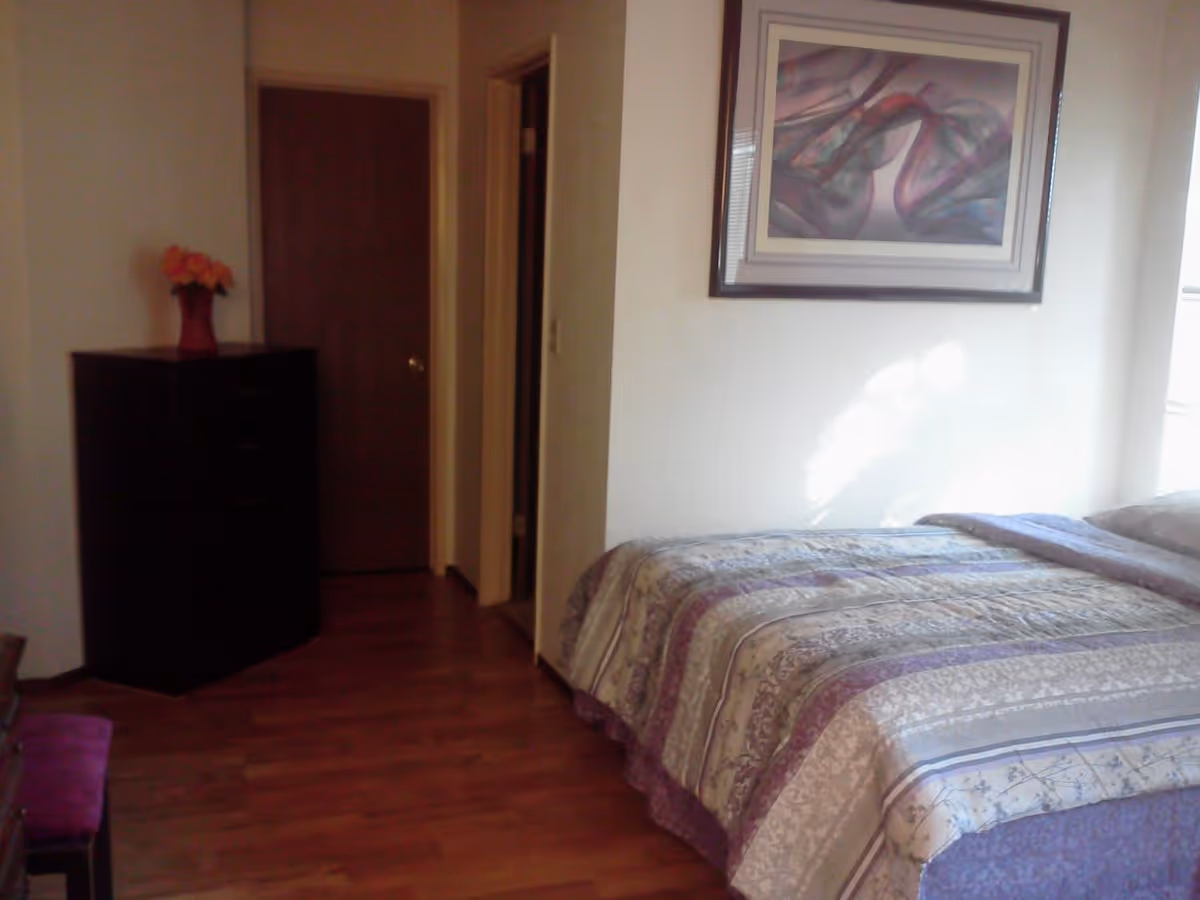 A bedroom with a bed covered in a patterned quilt, a framed abstract painting on the wall above the bed, a wooden dresser with a vase of flowers on top, and a wooden door leading to another room. The floor is wooden and there is natural light coming through a window.
