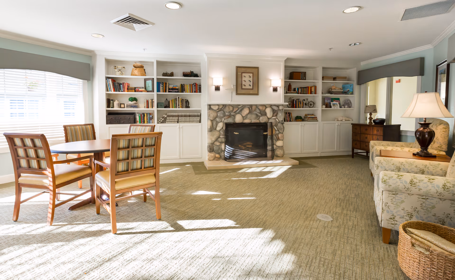 Sunlit common room with a round table and chairs, built-in bookshelves flanking a stone fireplace, and upholstered seating.