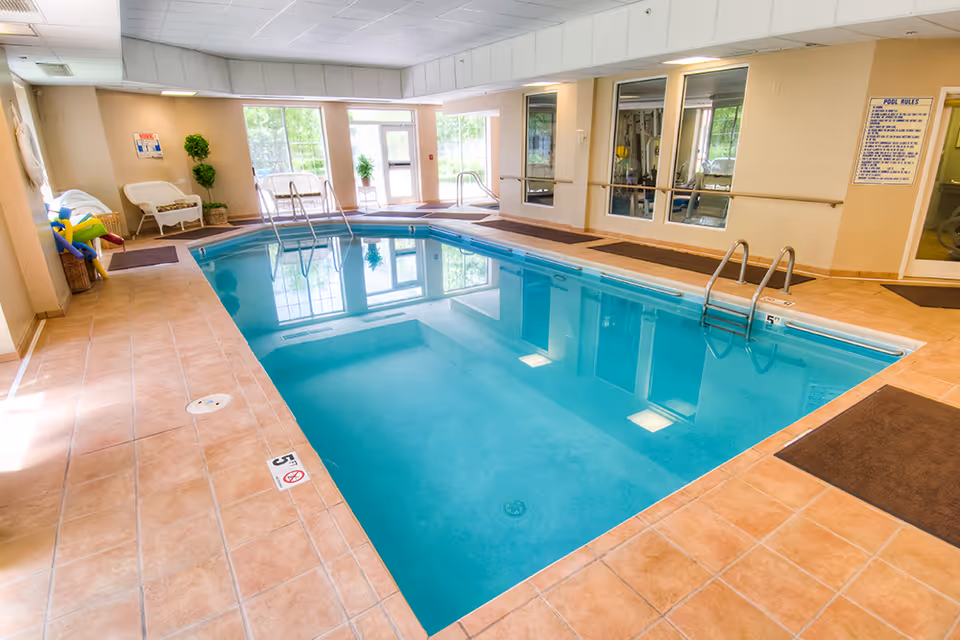Indoor swimming pool area with clear blue water, surrounded by beige tiled flooring. There are white chairs and potted plants near large windows letting in natural light. Pool rules are posted on the wall, and there are handrails for entering the pool.