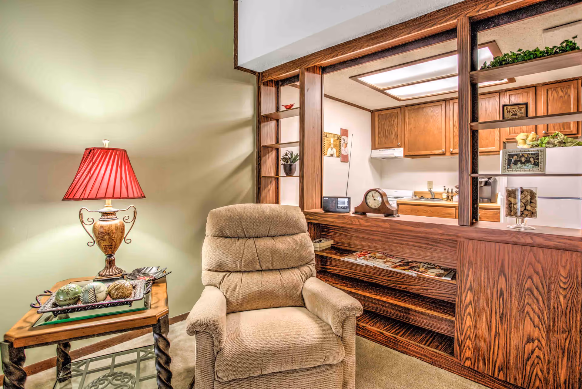 Cozy living room area with a recliner, side table and lamp facing a wood-paneled pass-through to a kitchen.