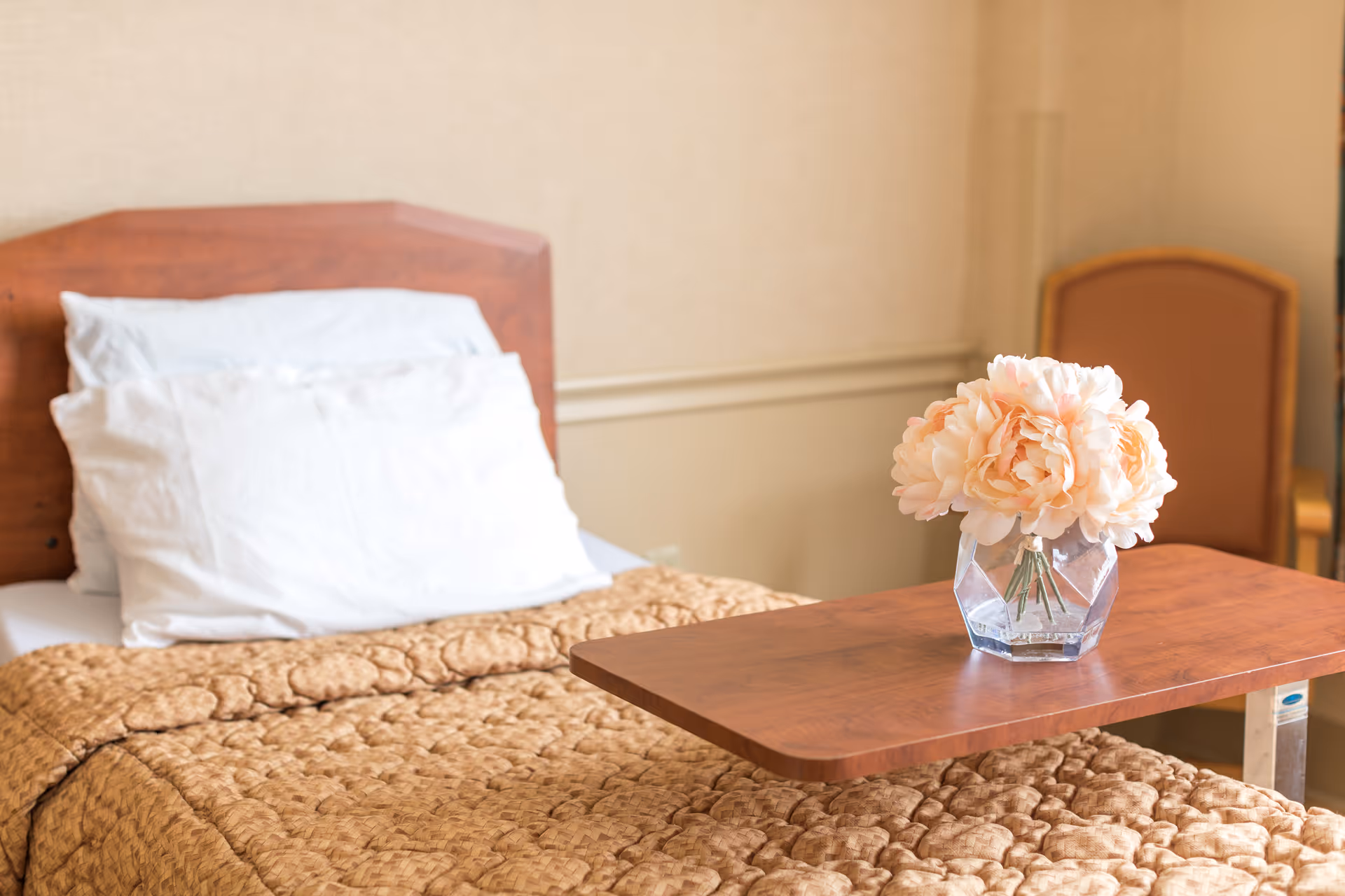 A single bed with white pillows and a brown quilted blanket. In front of the bed is a wooden overbed table holding a glass vase with light peach-colored flowers. In the background, there is a brown cushioned chair against a beige wall.