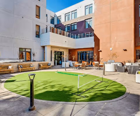 Sunlit enclosed courtyard with a round artificial turf putting-green area and outdoor seating surrounded by a multi-story assisted living building.
