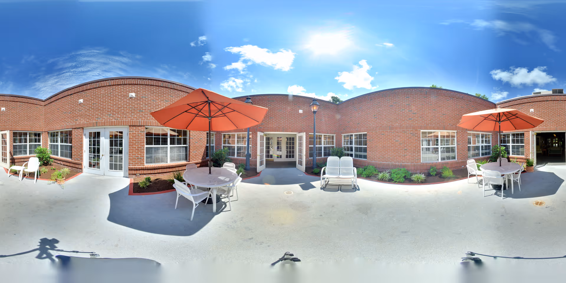Outdoor patio area at Oaks at Mt. Pleasant with round tables, white chairs, and orange umbrellas. The patio is surrounded by a brick building with multiple windows and doors. There are small garden beds with green plants along the building walls under a bright blue sky with some clouds.