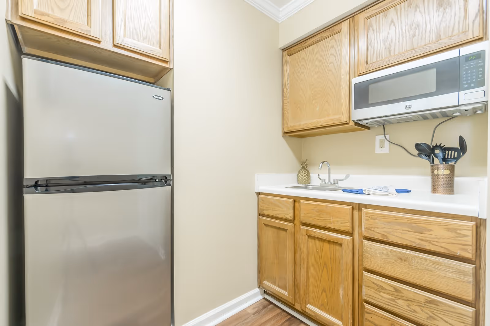 A small kitchen area featuring a stainless steel refrigerator, wooden cabinets, a white countertop with a sink, a microwave mounted above the counter, and a container holding kitchen utensils.
