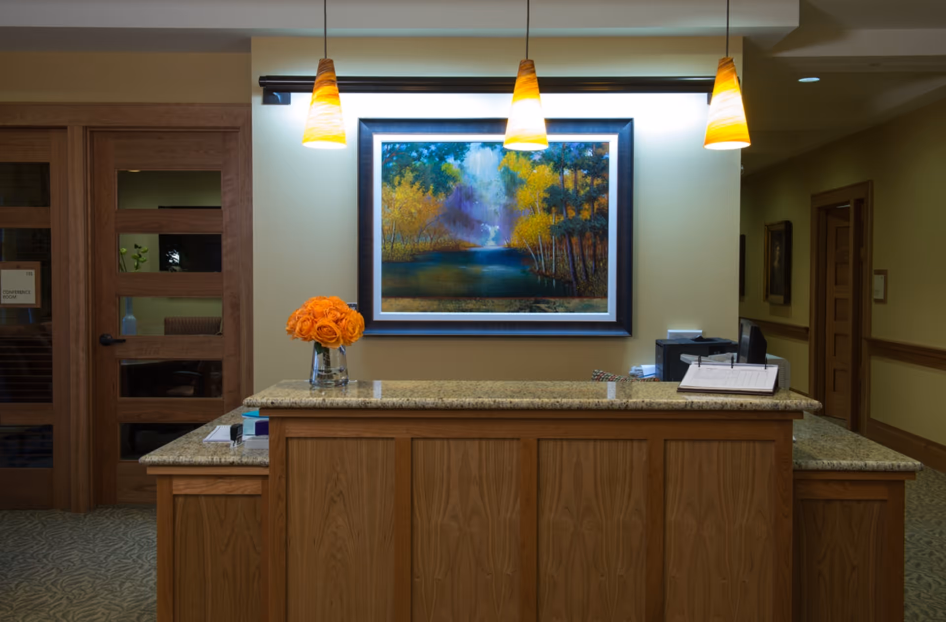 Reception desk area with a granite countertop and wooden paneling. A vase with orange flowers sits on the desk. Behind the desk is a wall with a large framed painting of a forest scene with a river, illuminated by three hanging pendant lights. To the left, there is a wooden door with glass panels labeled 'Conference Room'.
