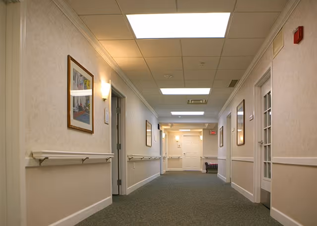 A well-lit hallway in a senior living facility with beige walls, carpeted floor, handrails on both sides, framed pictures on the walls, and several doors leading to different rooms. The ceiling has recessed lighting panels and a fire alarm is visible on the right wall.