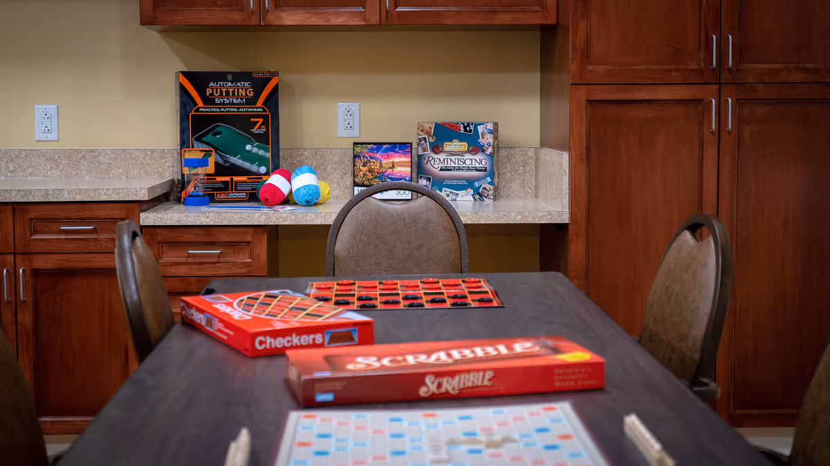 A room with a dark wooden table and four chairs. On the table are board games including Scrabble and Checkers. In the background, there are wooden cabinets and a countertop with additional games and colorful yarn balls placed on it.
