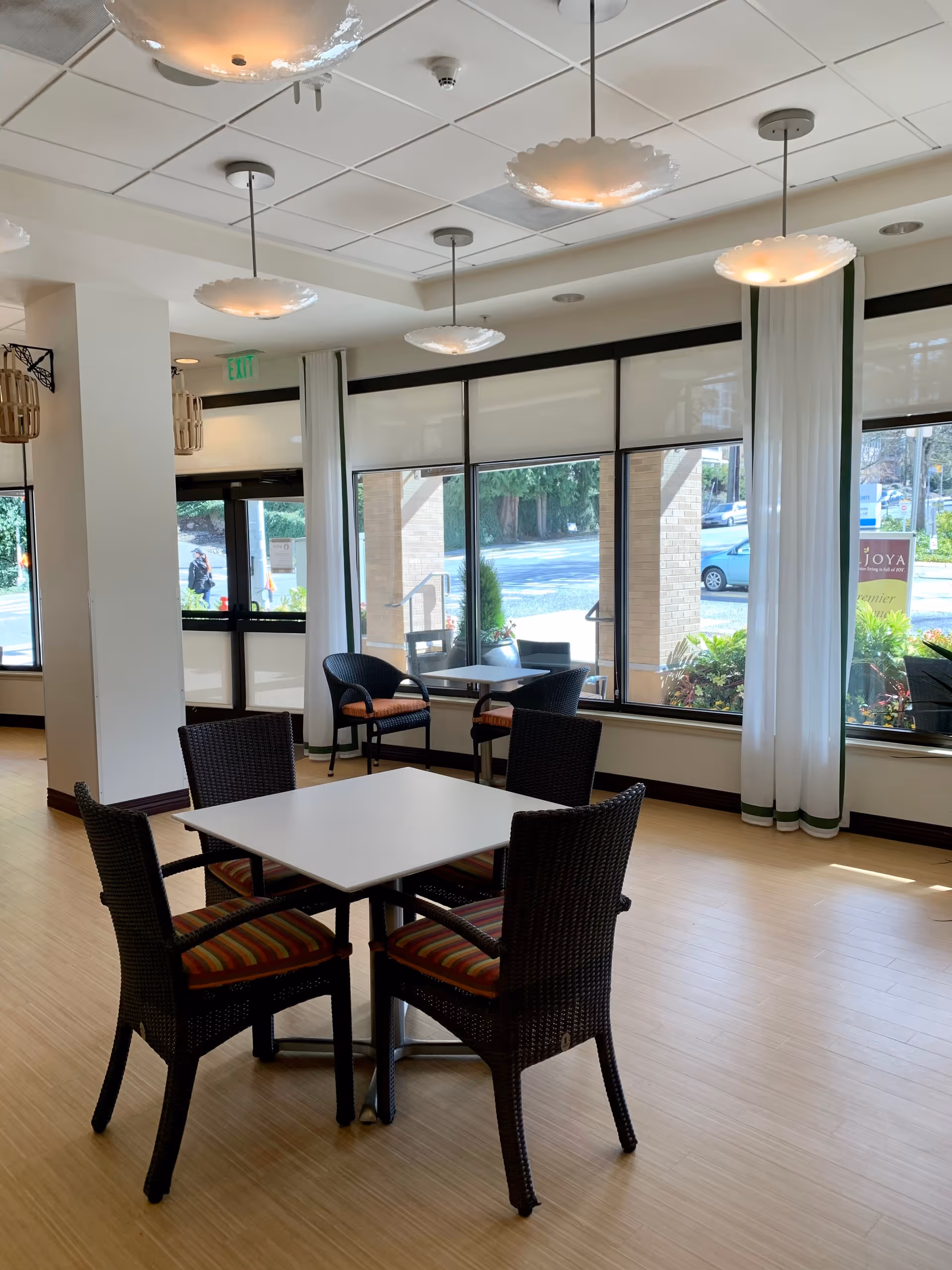 A bright and airy dining area with a square white table surrounded by four wicker chairs with striped cushions. Large windows with white curtains allow natural light to fill the room, and additional seating is visible near the windows. Modern ceiling lights hang above, and the floor is light wood.