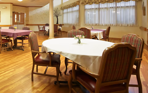 A dining room with several tables covered with tablecloths and small flower arrangements. The room has wooden floors, large windows with sheer curtains, and cushioned chairs with plaid upholstery.