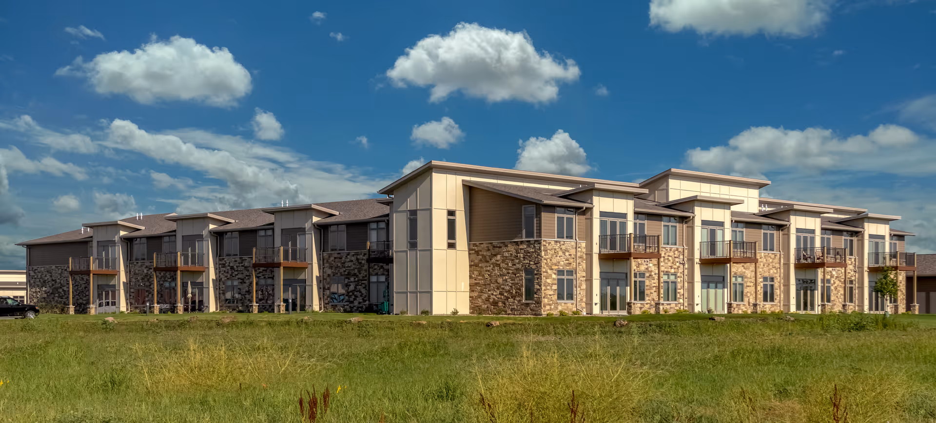Two-story modern senior living building with stone accents and balconies seen across a grassy foreground under a blue sky.