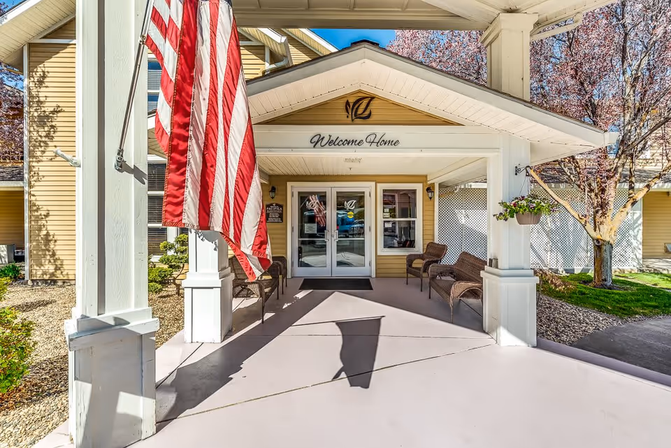 Entrance to Klamath Falls Senior Living facility with a covered porch, American flag hanging on the left, two wicker chairs on each side, and a sign above the door that reads 'Welcome Home'. Trees and landscaping are visible around the building.
