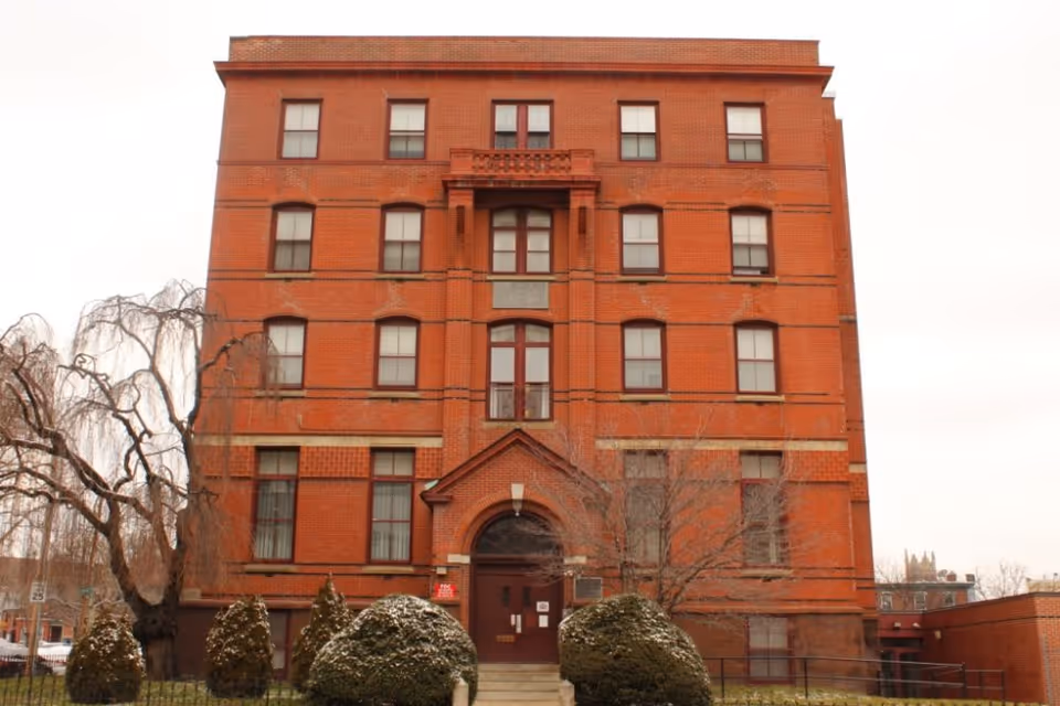 Front view of a red brick multi-story building with a central arched entrance, rows of windows, and shrubs in the foreground.