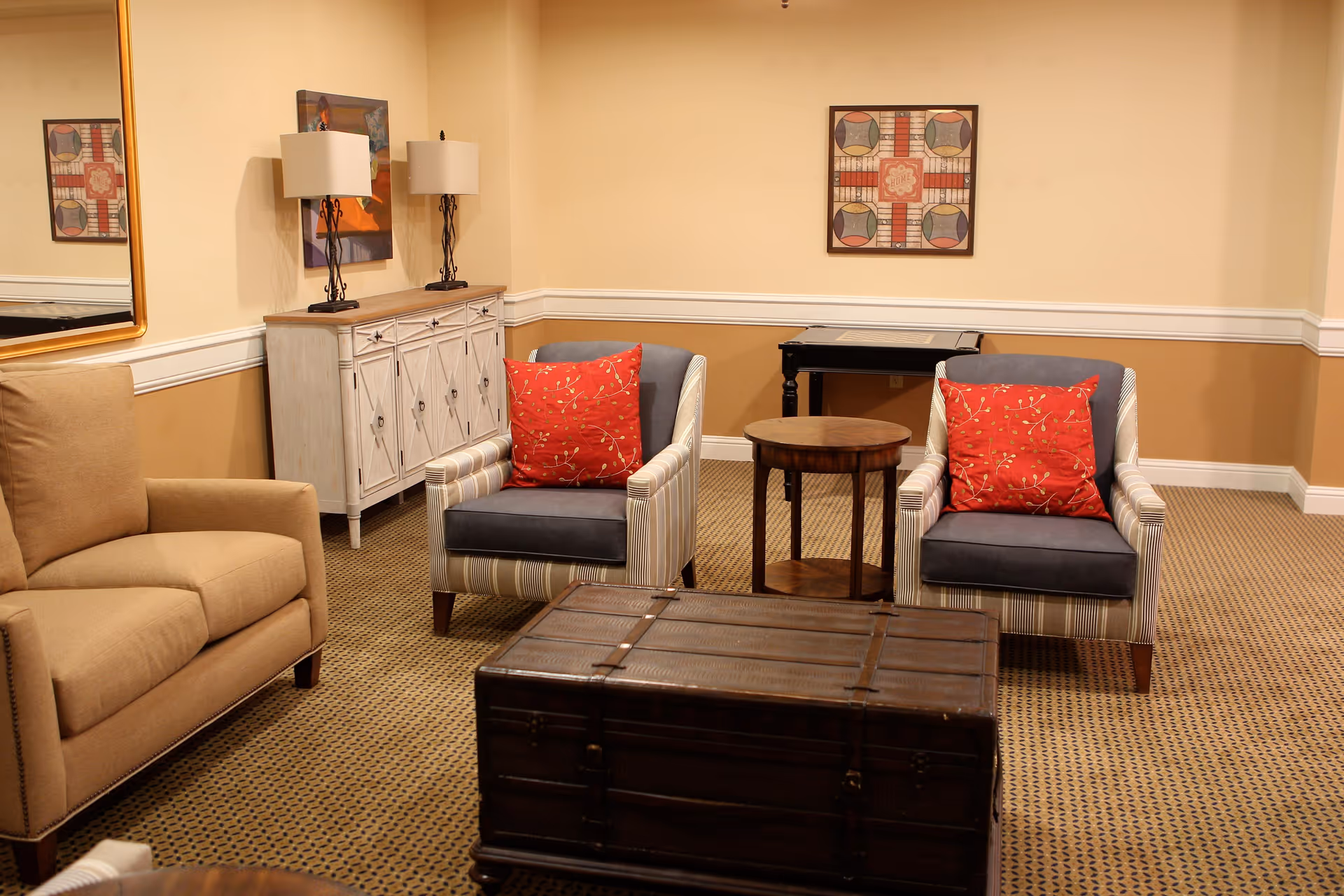 A cozy living room area with two striped armchairs each with a red decorative pillow, a beige armchair, a wooden chest used as a coffee table, a small round wooden side table, a white cabinet with two lamps on top, and framed artwork on the walls.