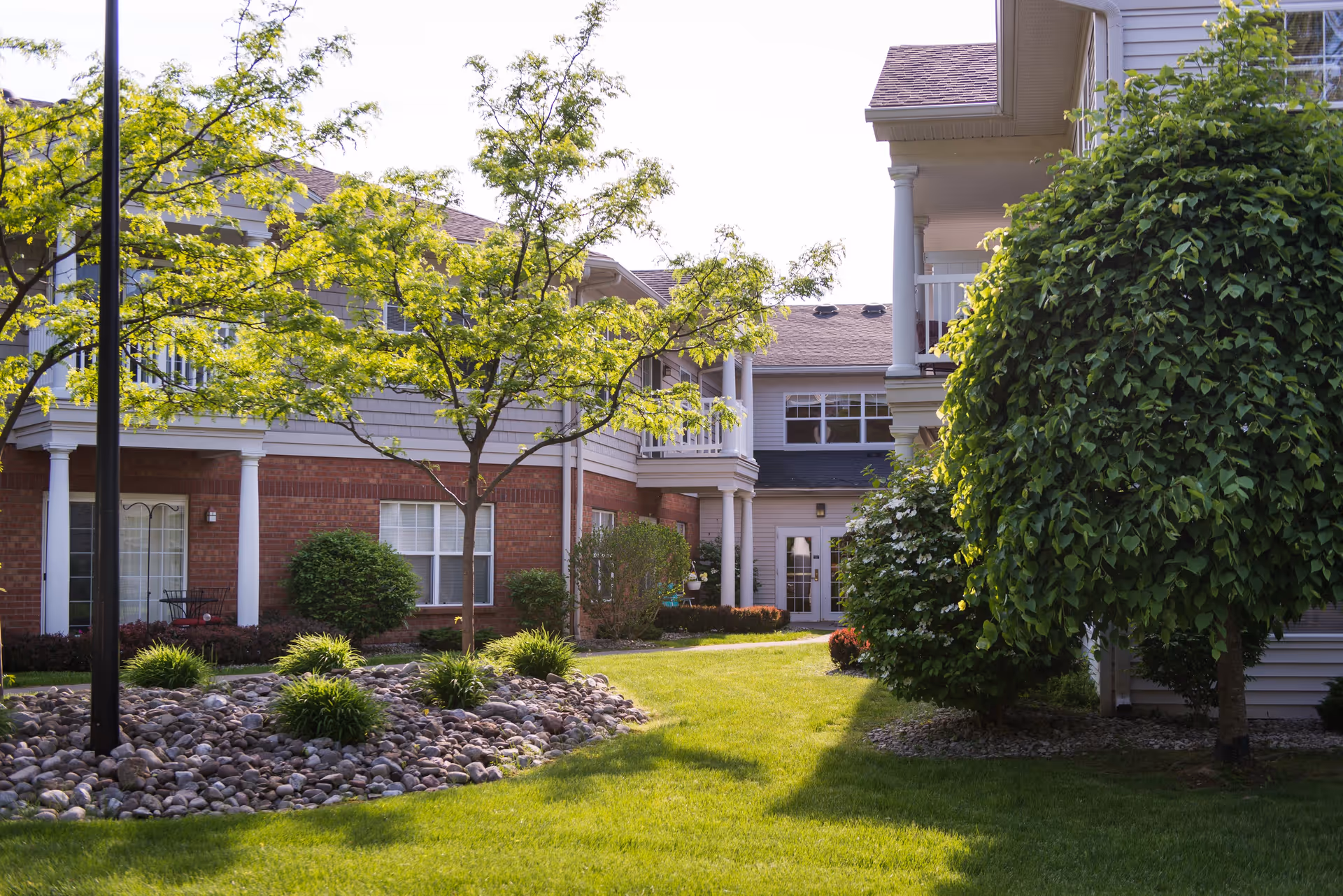 Outdoor view of a senior living facility courtyard with green grass, trees, bushes, and a pathway. The building has brick and siding exterior with white columns and balconies.