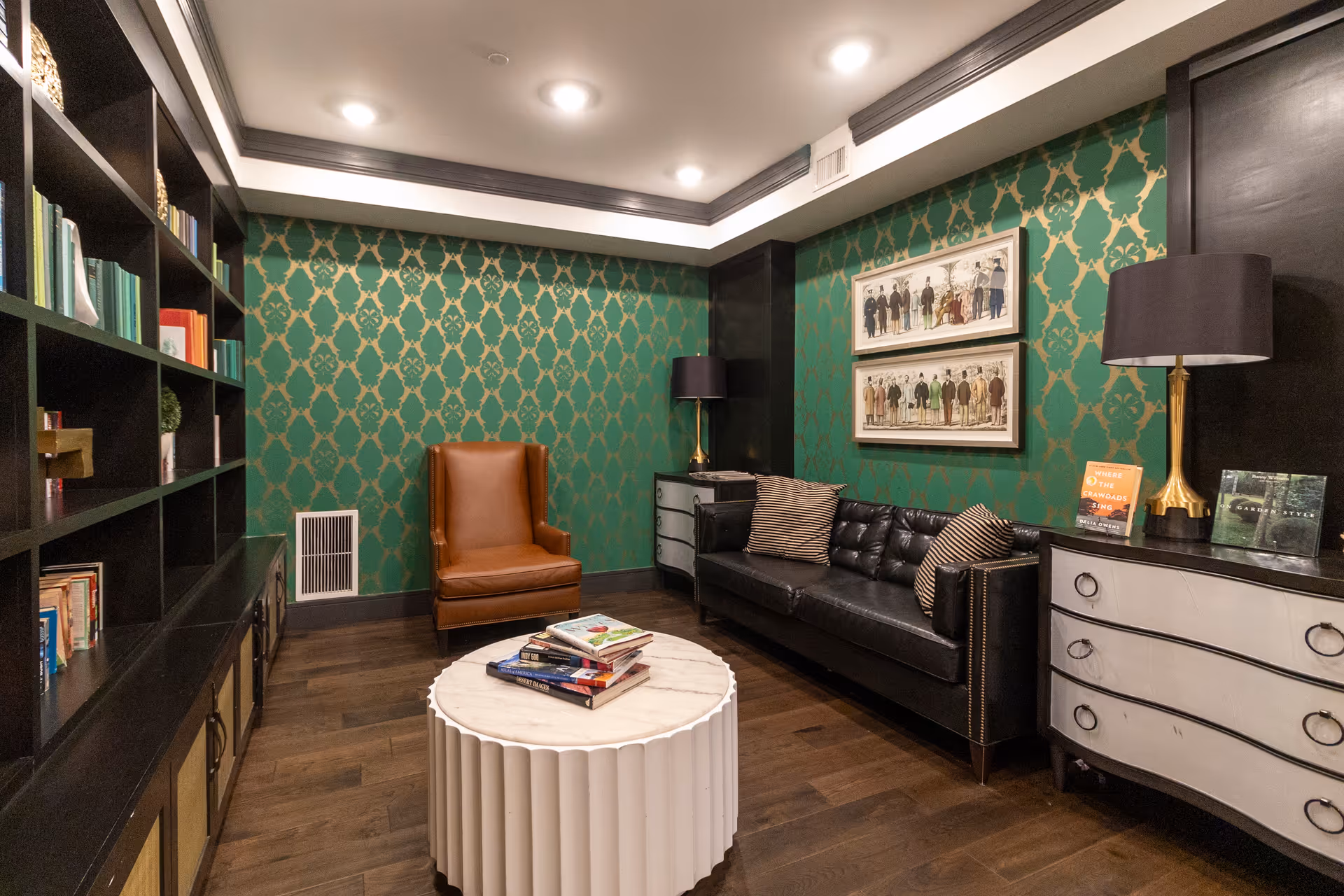 Cozy lounge with green patterned wallpaper, black leather sofa, brown armchair, bookshelves and round white coffee table.